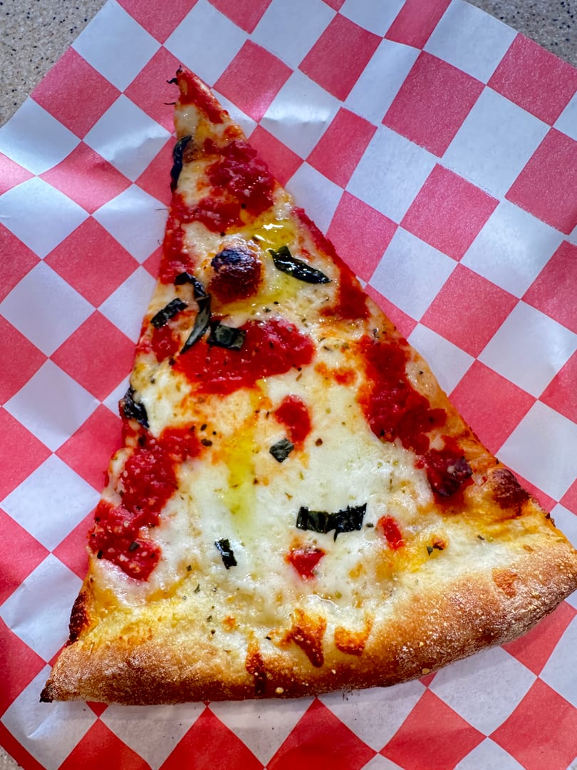 A slice of pizza with red sauce, melted cheese, and basil on red and white checkered paper.