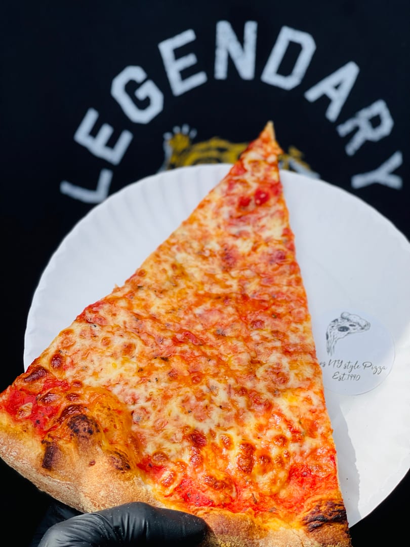 A hand in a black glove holds a cheesy pizza slice on a white plate, with a "Legendary" shirt in the background.