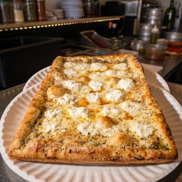 A rectangular white pizza with dollops of ricotta cheese and herbs on a paper plate.