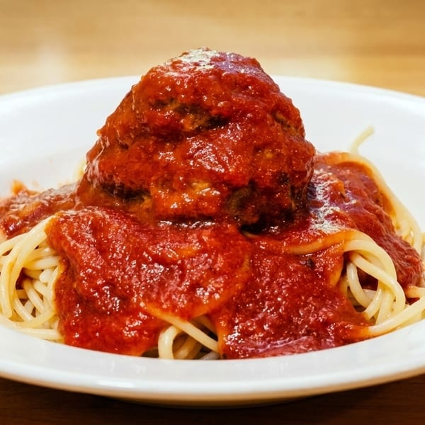 Spaghetti and a large meatball covered in rich red marinara sauce on a white plate.