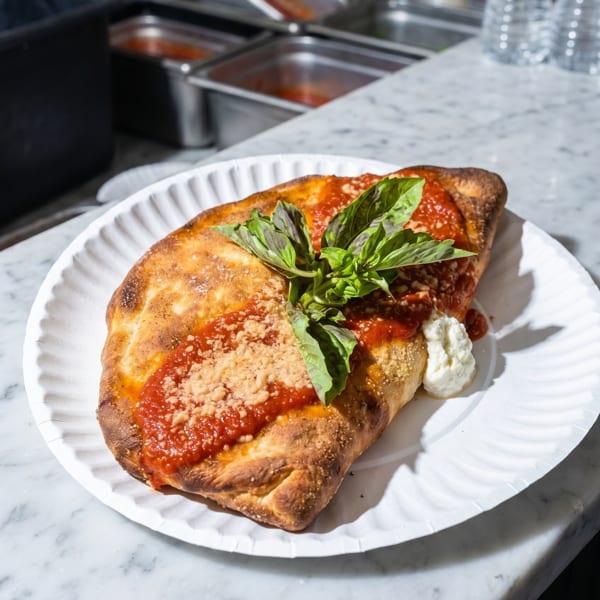 A calzone topped with red sauce, parmesan, fresh basil, and a dollop of ricotta on a paper plate.